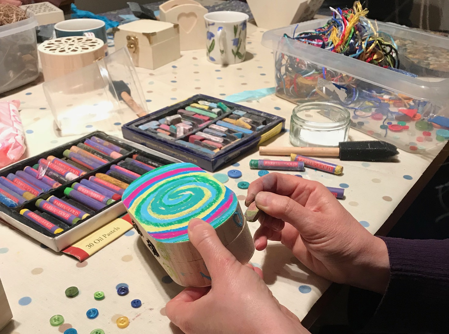 A person decorates a box with colorful spiral patterns using oil pastels, surrounded by art supplies on a table.