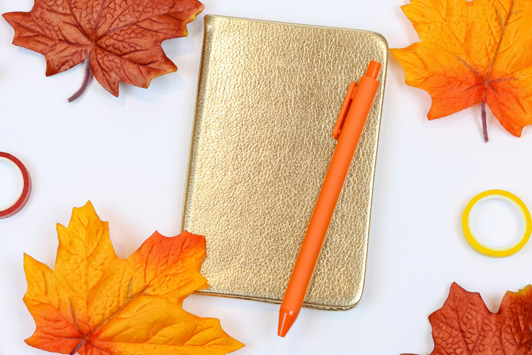 A gold notebook and pen surrounded by orange and red autumn leaves.