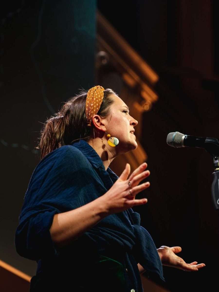 Looking up at a woman performing on a stage in front of a microphone wearing a yellow headband and adopting a dramatic pose.
