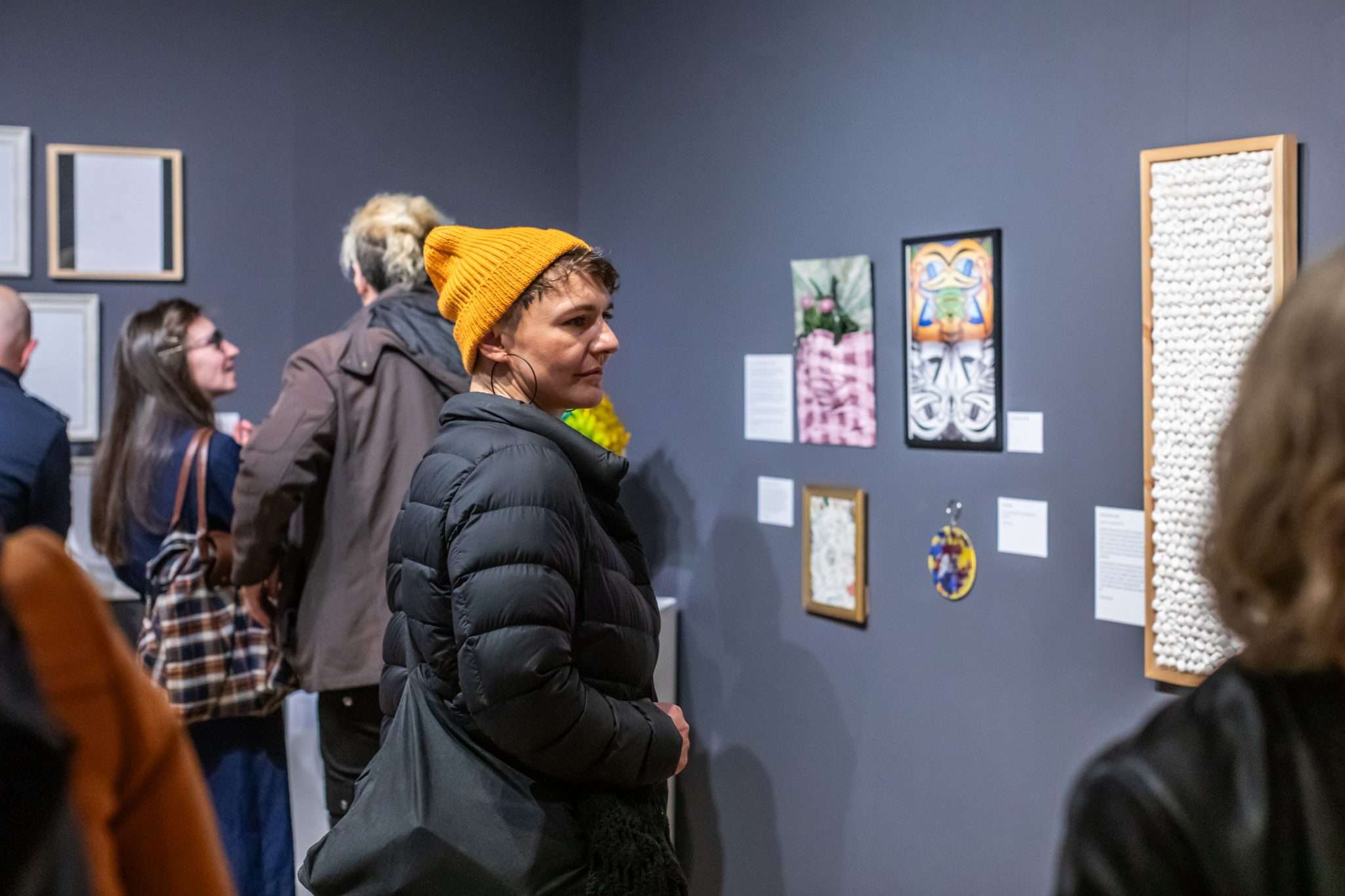 Woman in a yellow woolen hat views an exhibition in art gallery, surrounded by other people.