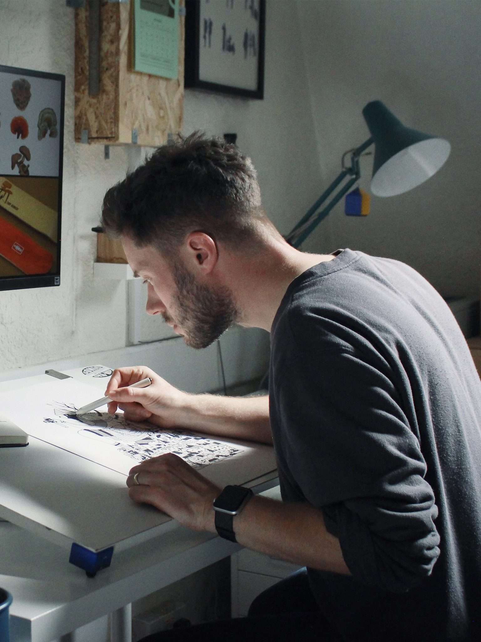 Man draws in a studio on a large sheet of paper with a monitor in front of him and stationery in a box at his side.