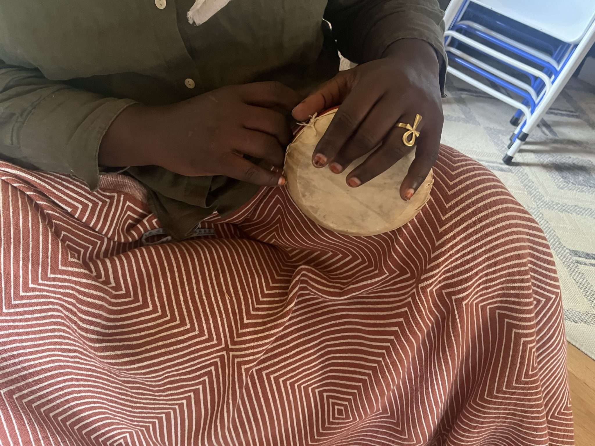 A black woman with painted nails plays a small drum while sitting on the floor under a blanket.